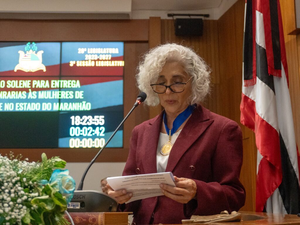 Dra. Maria dos Remédios Branco discursa na tribuna da Assembleia Legislativa do Maranhão durante a cerimônia de entrega da Medalha de Mérito Legislativo Jackson Lago, usando a medalha no peito e segurando o discurso impresso.