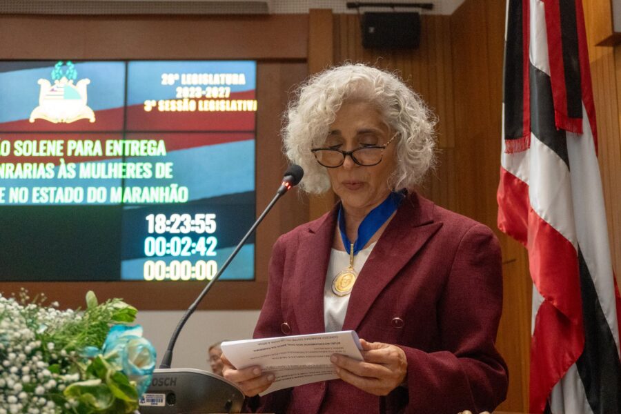 Dra. Maria dos Remédios Branco discursa na tribuna da Assembleia Legislativa do Maranhão durante a cerimônia de entrega da Medalha de Mérito Legislativo Jackson Lago, usando a medalha no peito e segurando o discurso impresso.