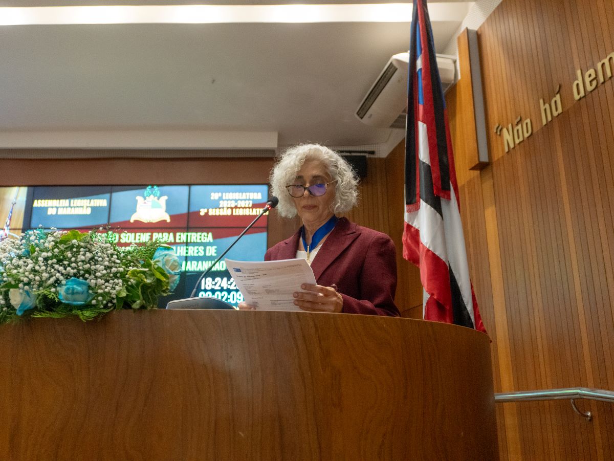 Maria dos Remédios, com medalha no peito, faz discurso na tribuna da Assembleia Legislativa do Maranhão, ao lado da bandeira do estado